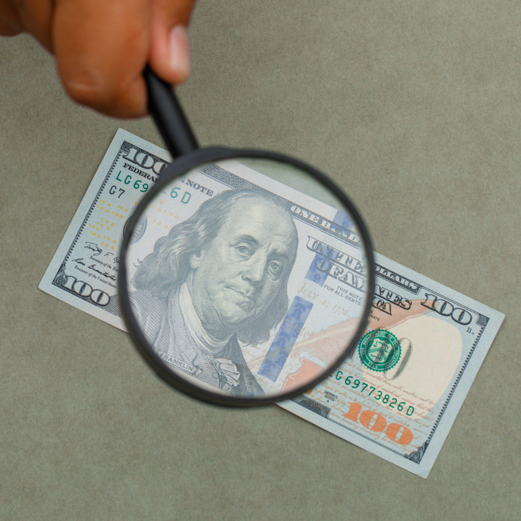 Hand holding a magnifying glass over a $100 bill on a beige background