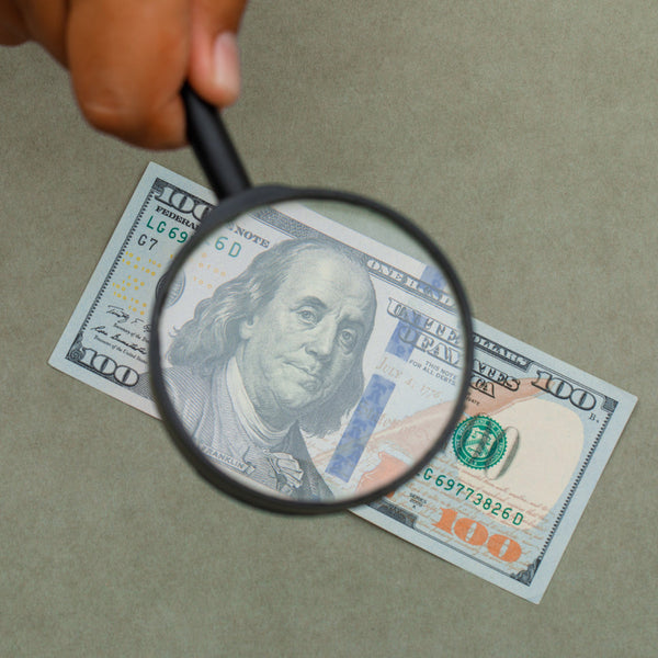 Hand holding a magnifying glass over a $100 bill on a beige background